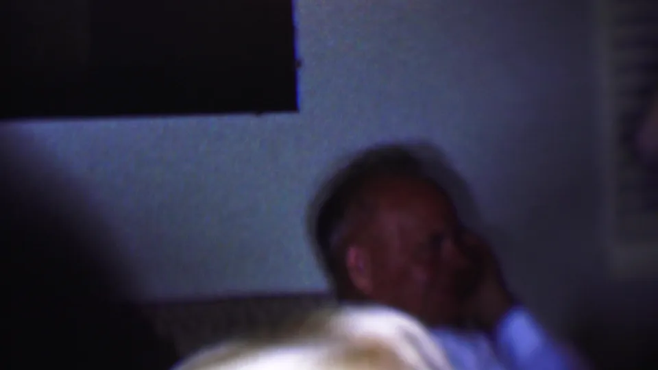 A seated adult man is partly obscured by shadow in a dim Phoenix interior, framed against a pale wall and dark rectangular opening.