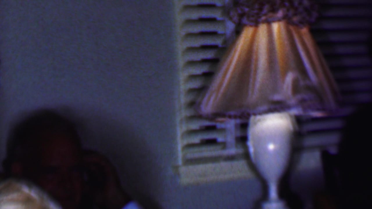 1956 Phoenix home-movie frame showing a glowing pleated table lamp near a shuttered window, with seated adults partly visible in a dim private interior.