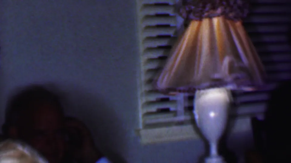 1956 Phoenix home-movie frame showing a glowing pleated table lamp near a shuttered window, with seated adults partly visible in a dim private interior.
