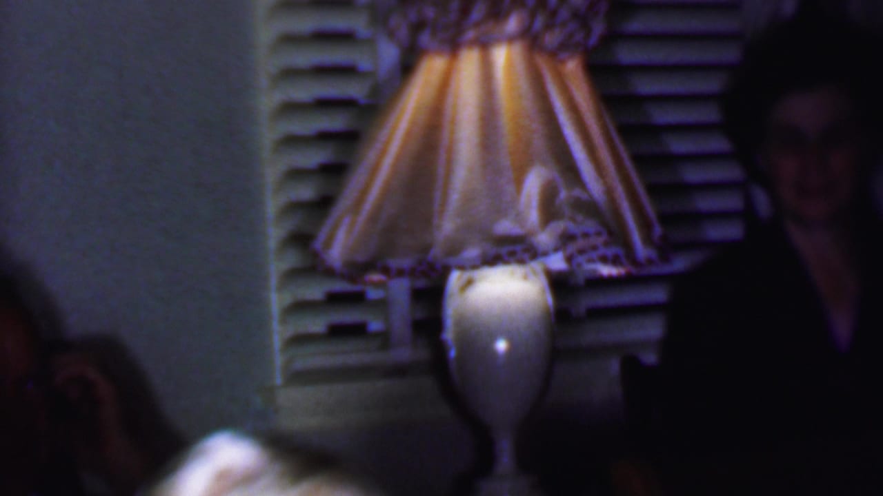 1956 Phoenix home-movie frame with a lit table lamp, shuttered window, and partially visible adults seated or standing in a dark home interior.