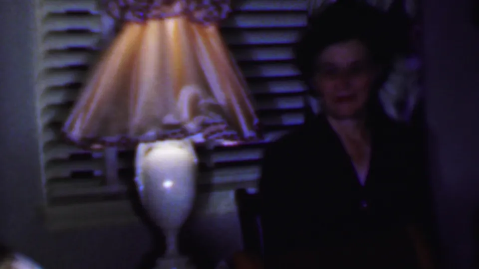 1956 Phoenix home-movie frame showing a woman in a dark dress standing beside a lit table lamp and shuttered window during an indoor gathering.