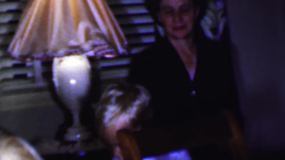 1956 Phoenix home-movie frame with a standing woman near a glowing fringed lamp and a partially visible seated person in the foreground of a private room.