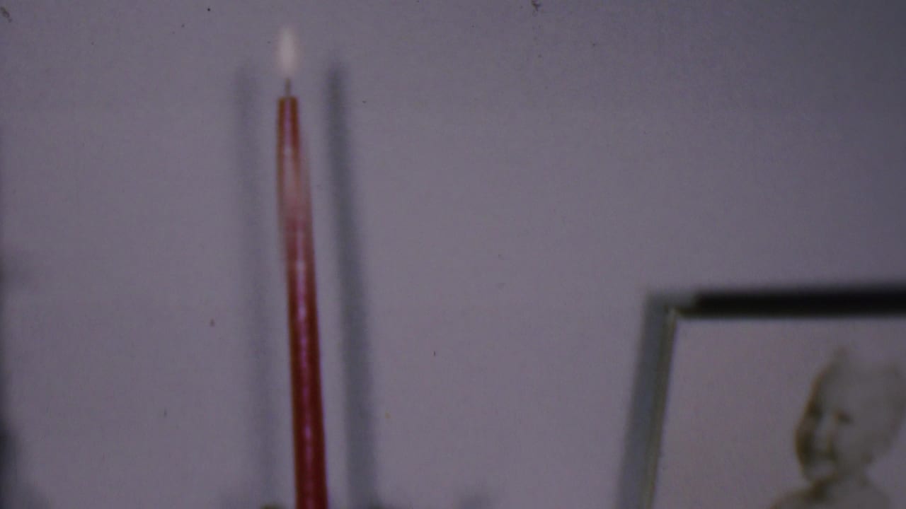 Phoenix, Arizona 1956 home-movie frame showing a lit red taper candle on or near a wall display, with a pale wall and blurred framed picture in the background.