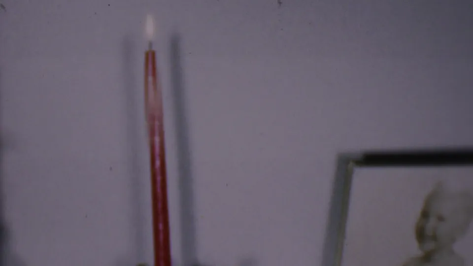 Phoenix, Arizona 1956 home-movie frame showing a lit red taper candle on or near a wall display, with a pale wall and blurred framed picture in the background.