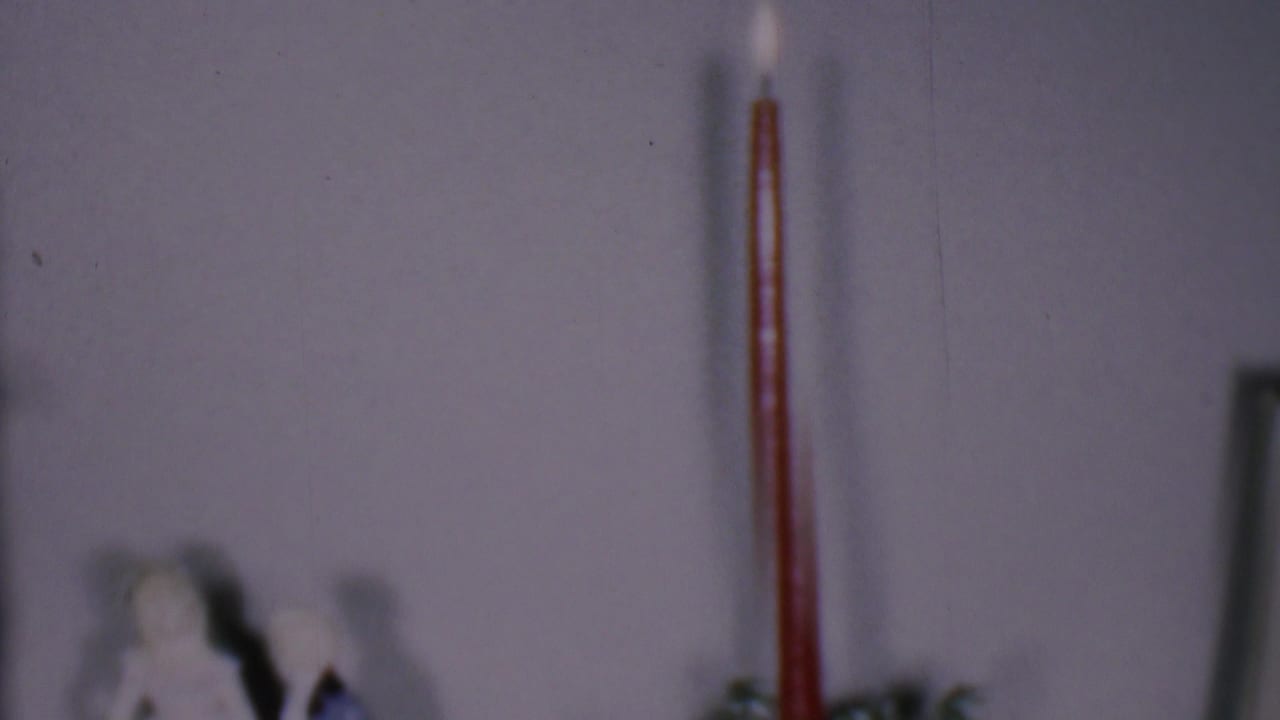 Phoenix, Arizona 1956 home-movie frame showing a lit red candle above a soft-focus holiday display with a small figurine and pale wall behind it.