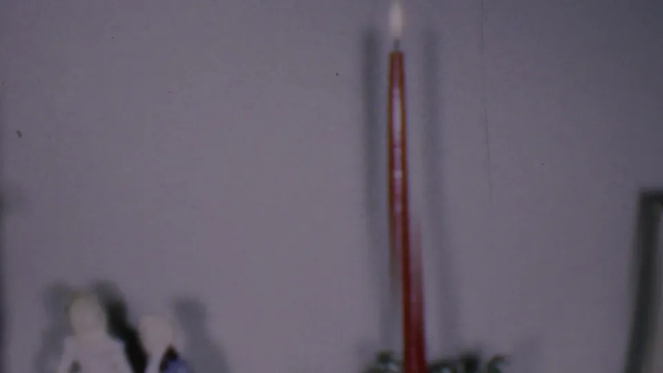 Phoenix, Arizona 1956 home-movie frame showing a lit red candle above a soft-focus holiday display with a small figurine and pale wall behind it.