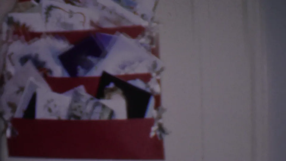 Phoenix, Arizona 1956 home-movie frame showing a red wall shelf crowded with holiday cards and trim beneath a red taper candle in a private interior.