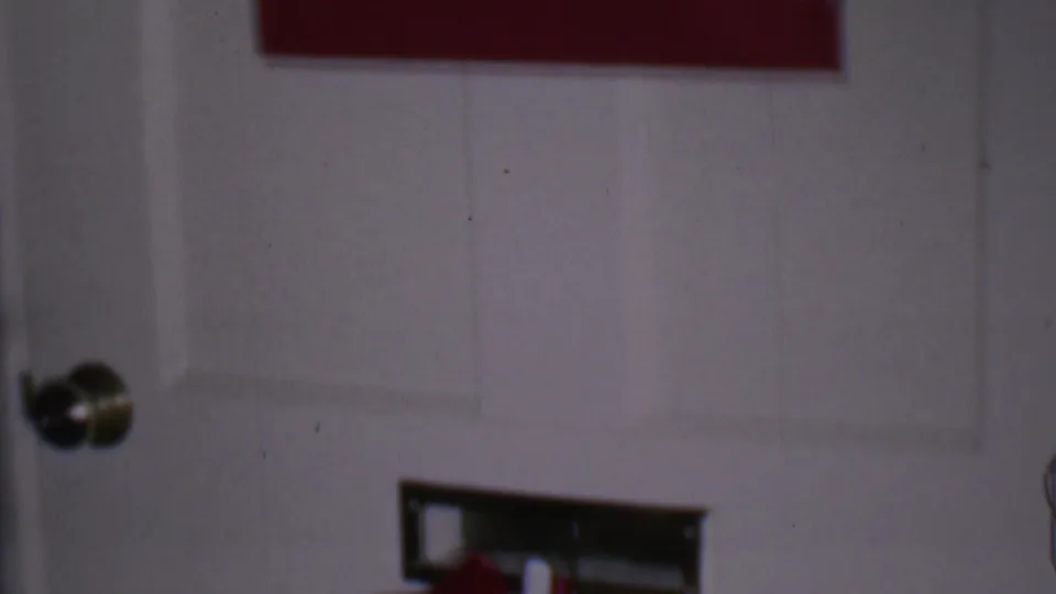 1956 Phoenix home-movie frame showing the lower portion of an interior door, a brass-colored doorknob, and the edge of a red-accented holiday card display.