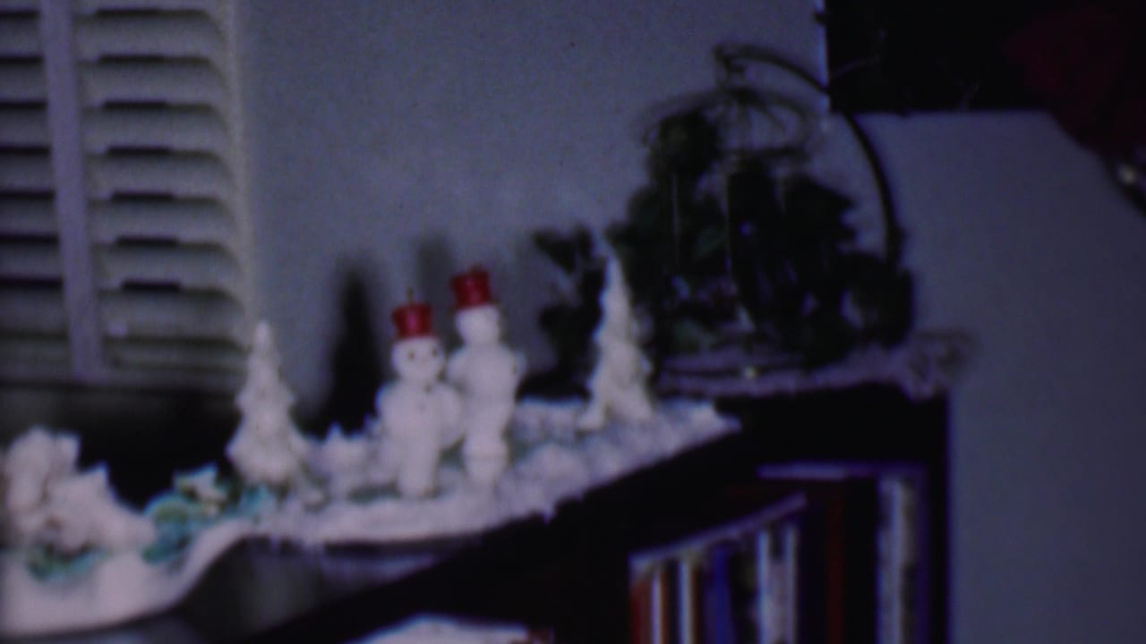 Soft 8mm frame from a 1956 Phoenix home movie showing small snowmen in red hats, white tabletop Christmas trees, window shutters, and indoor holiday display pieces.