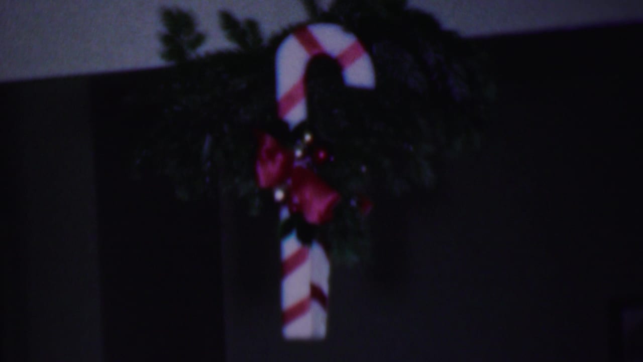 1956 Phoenix home-movie frame showing a large red-and-white candy cane ornament hanging with greenery and red ribbon in a dim home interior.