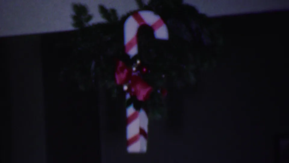 1956 Phoenix home-movie frame showing a large red-and-white candy cane ornament hanging with greenery and red ribbon in a dim home interior.