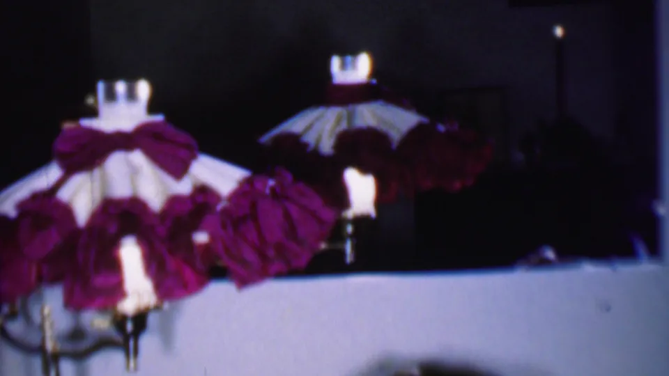 1956 Phoenix 8mm home-movie frame showing decorative lamps or candle-style holiday pieces with red ruffled shades arranged along an indoor ledge beneath a dark room background.