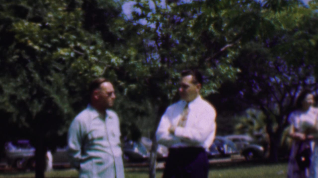 Two adult men stand in conversation in a Dallas park-like setting in 1956, with leafy trees and mid-century cars visible behind them.