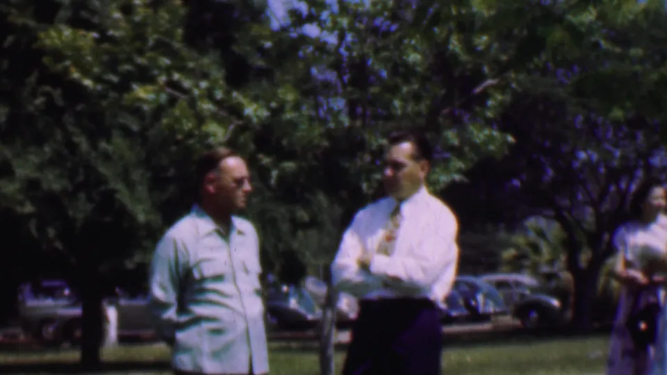 The two men remain in conversation in the 1956 Dallas outdoor scene, with a woman partly visible behind them and cars parked along the background.