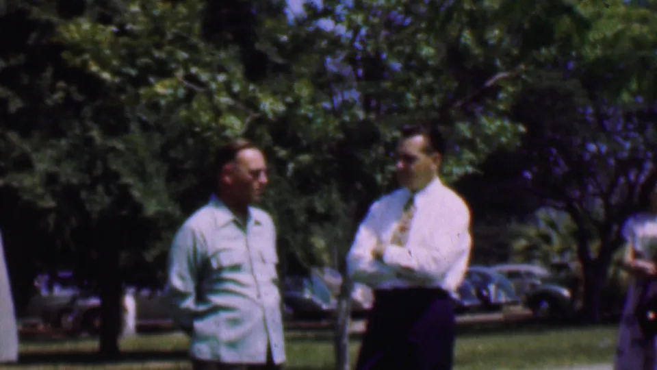 Two men stand near a walkway and grass in the 1956 Dallas home-movie scene as a woman in a light top and patterned skirt appears at the right edge.