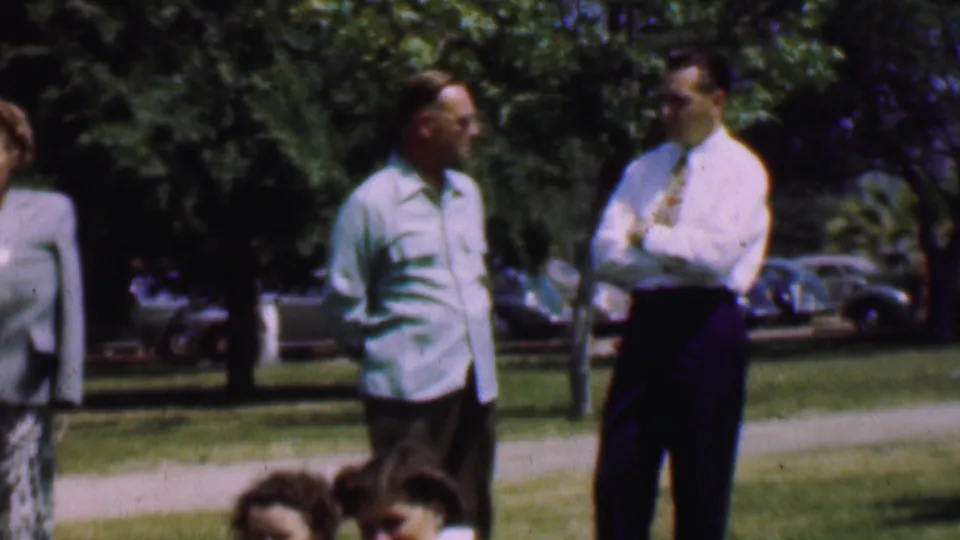 The men continue their outdoor conversation in the Dallas 1956 clip, with a walkway, trees, parked cars, and partially seen adults around the frame edges.
