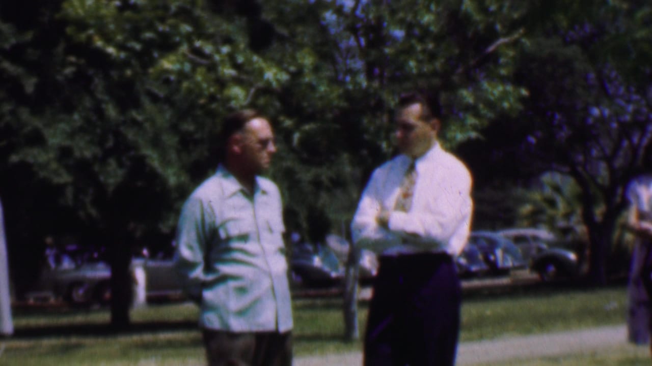 In Dallas, Texas, 1956, two adult men stand outdoors on a grassy area, one in a light shirt and the other in a white shirt with tie, with leafy trees and parked mid-century cars in the background.