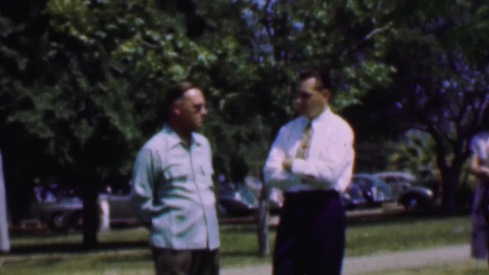 In Dallas, Texas, 1956, two adult men stand outdoors on a grassy area, one in a light shirt and the other in a white shirt with tie, with leafy trees and parked mid-century cars in the background.