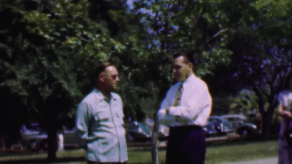 Two adult men face one another in a Dallas, Texas, 1956 outdoor setting, framed by grass, deep green trees, and several parked mid-century cars behind them.