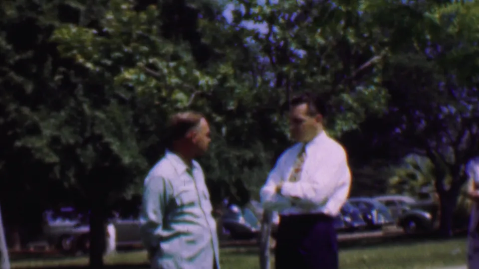 At the end of the clip, the two men remain outdoors in the 1956 Dallas scene, with one turned toward the other and classic cars visible beyond the grassy, tree-lined area.