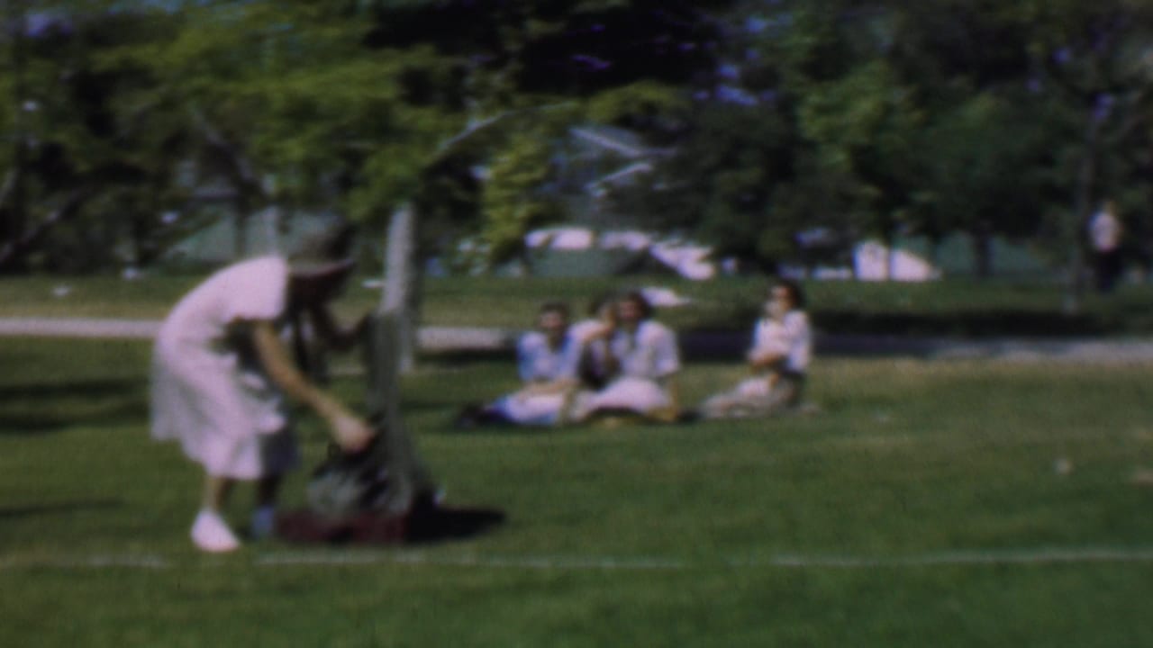 In this 1956 Dallas home-movie frame, a woman in a light dress and hat bends toward shoes and a dark jacket on the grass, with several women seated farther back in a sunny park.