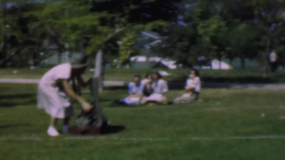 In this 1956 Dallas home-movie frame, a woman in a light dress and hat bends toward shoes and a dark jacket on the grass, with several women seated farther back in a sunny park.