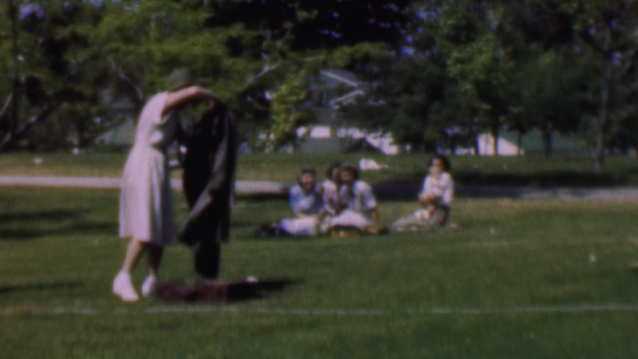 Woman lifts a dark jacket over her head on a grassy lawn with seated women and trees behind her.