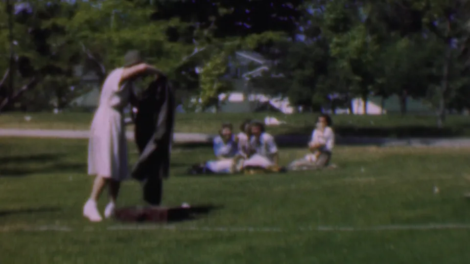 In the Dallas park setting, the woman raises a dark jacket or coat above her head while standing on the grass, with women seated in summer dresses in the background.