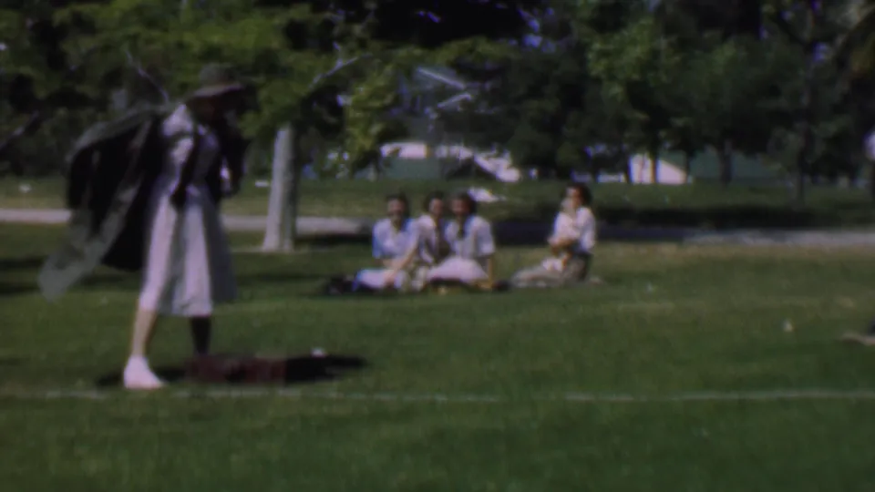 The woman stands in profile on the park lawn with a dark jacket partly around her shoulders, while a small group of women sits behind her under trees.