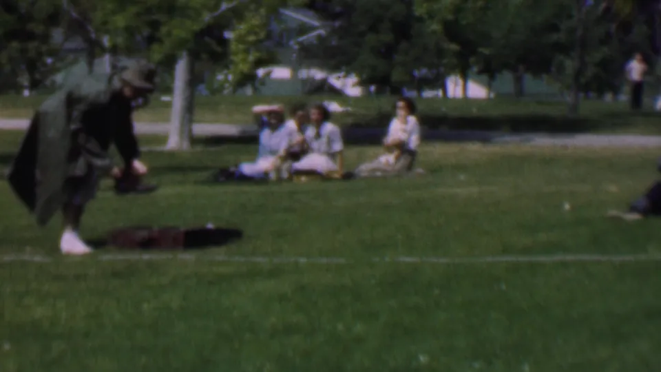 At the end of the clip, the woman bends forward with the dark jacket draped around her, still near shoes on the grass, while the seated group remains visible in the Dallas park scene.