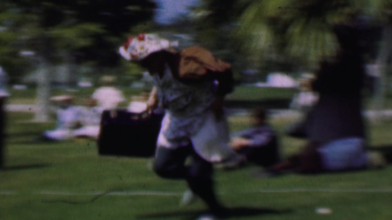A woman in a hat moves across a park lawn carrying a suitcase.