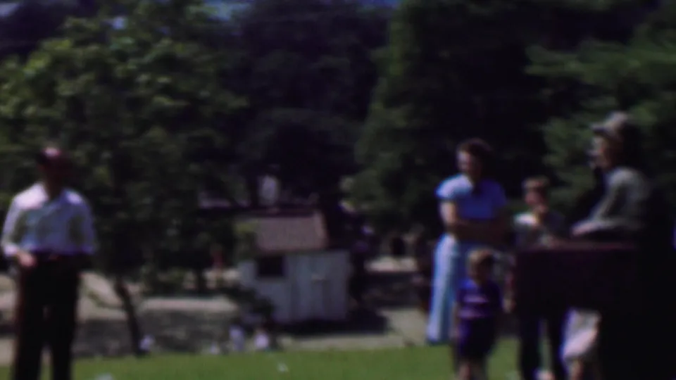 1956 Dallas 8mm frame showing a man standing at the edge of a lawn with groups of people farther back near a path and trees in a public park.