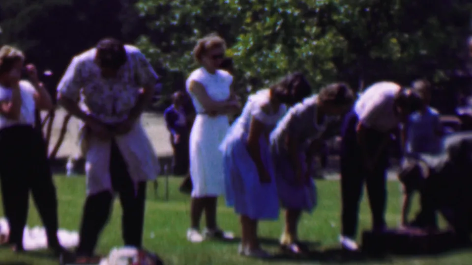 1956 Dallas home-movie frame showing adults gathered on a sunlit park lawn, with several people bending over clothing or suitcases while others stand nearby in dresses and casual wear.