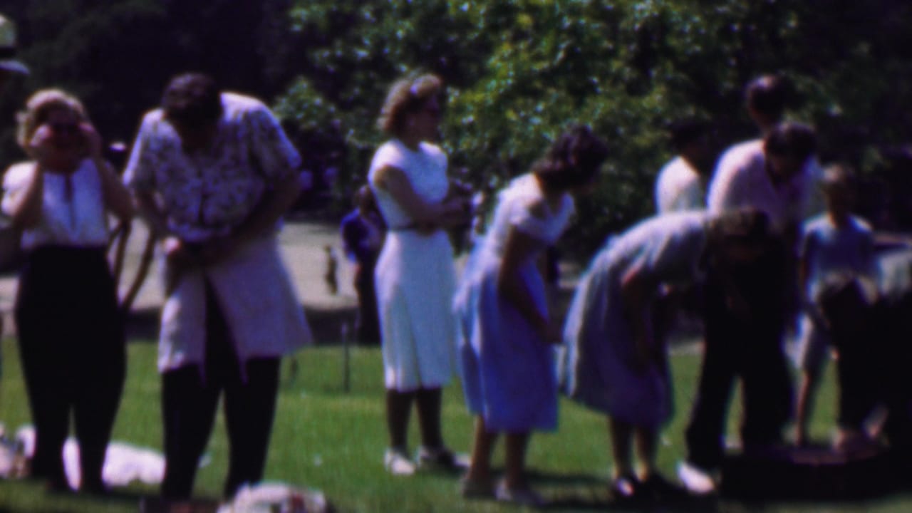1956 Dallas home-movie frame showing adults in mid-century clothing gathered on a park lawn, with trees behind them and several people looking down toward a blanket or belongings.