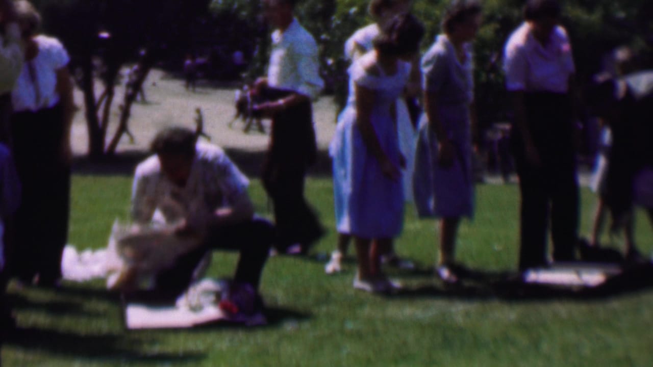 A group stands on a sunlit lawn while one person kneels near a blanket and small items.