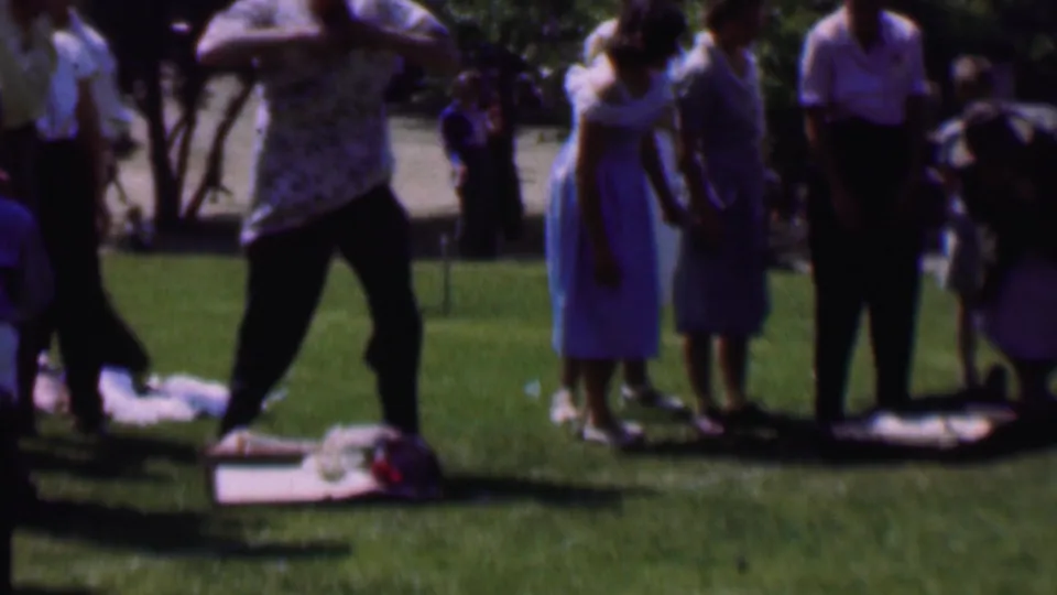 1956 Dallas home-movie frame showing a man in a patterned shirt moving beside a blanket on the lawn, with women in dresses and skirts standing nearby in the park.