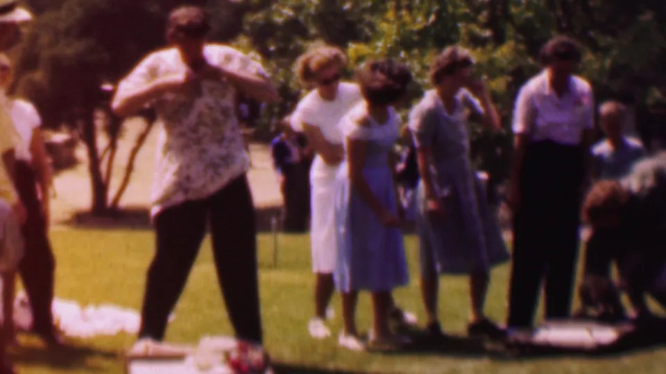 1956 Dallas park footage frame showing adults in mid-century clothing gathered on grass, with a man in a patterned shirt adjusting his clothing near a blanket and trees behind the group.