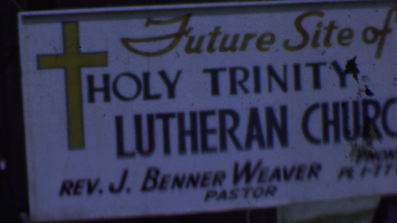 1956 Dallas home-movie frame showing a white future-site sign for Holy Trinity Lutheran Church, with a yellow cross and Rev. J. Benner Weaver named as pastor.