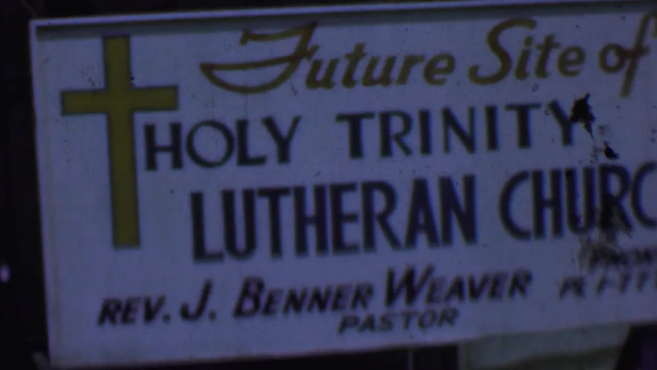 1956 Dallas home-movie frame showing a white future-site sign for Holy Trinity Lutheran Church, with a yellow cross and Rev. J. Benner Weaver named as pastor.