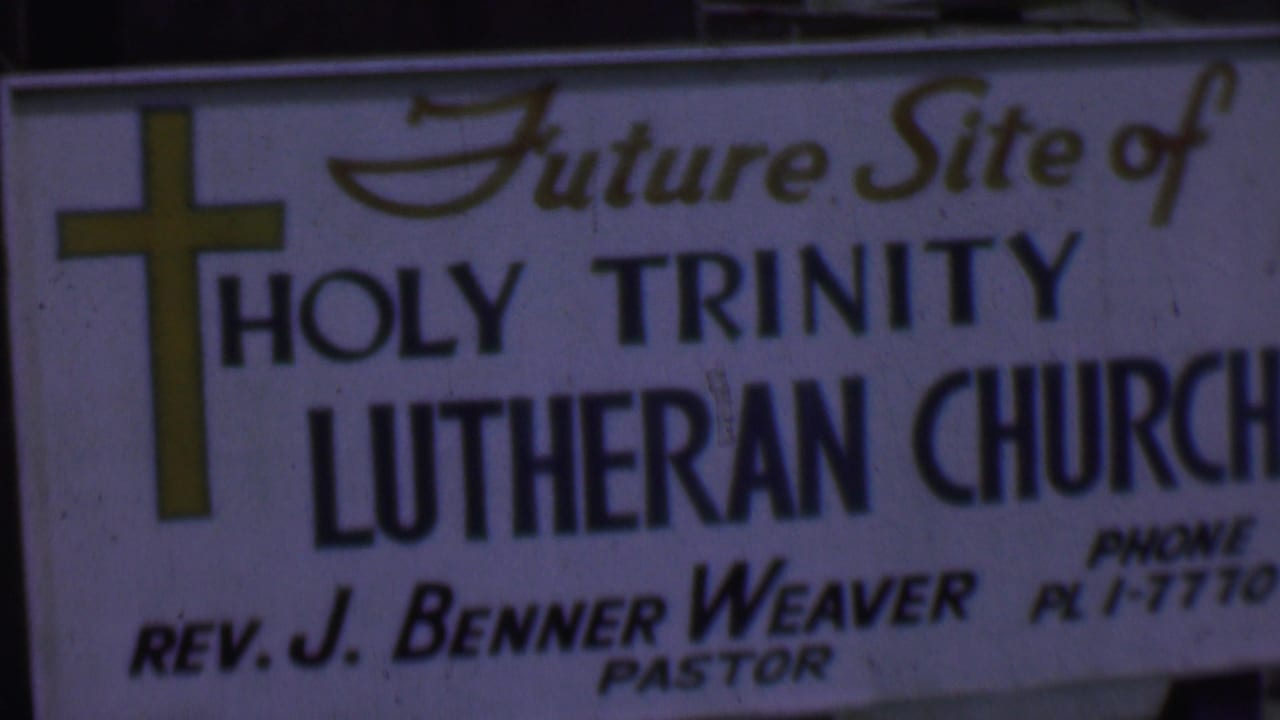 Close 1956 Dallas frame of the Holy Trinity Lutheran Church site sign, showing the yellow cross, large Lutheran Church lettering, and partially readable phone and pastor details.