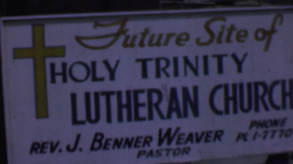 Close 1956 Dallas frame of the Holy Trinity Lutheran Church site sign, showing the yellow cross, large Lutheran Church lettering, and partially readable phone and pastor details.