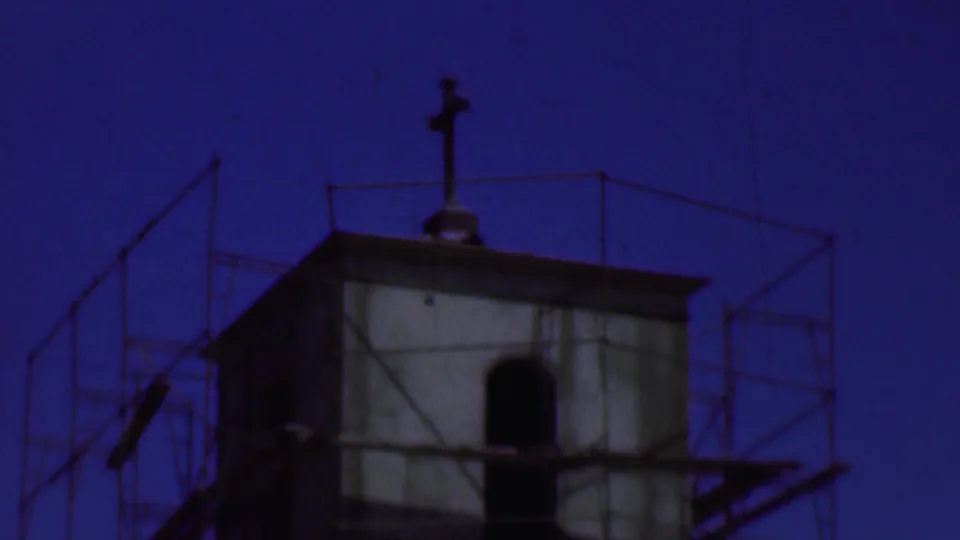 1956 Dallas frame showing the Holy Trinity Lutheran Church tower under construction, wrapped in scaffolding with a rooftop cross silhouetted against a deep blue twilight sky.