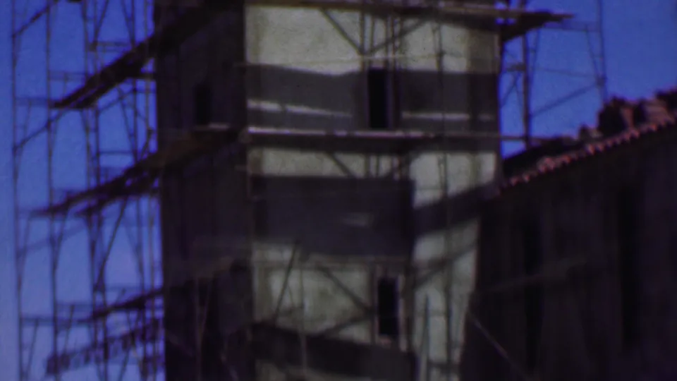 1956 Dallas 8mm still showing the lower portion of a scaffolded concrete tower beside a tiled roofline and open sky.