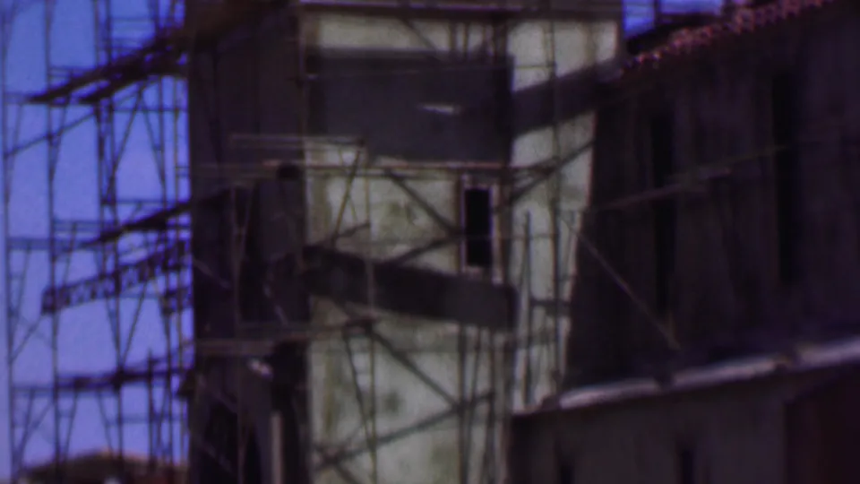 1956 Dallas frame showing the scaffolded tower from a slightly lower angle, with roof tiles, shadowed openings, and saturated blue sky.