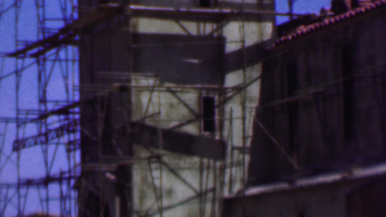 1956 Dallas 8mm frame showing a scaffolded building facade with a pale tower-like section, dark openings, and adjacent roofline under blue sky.