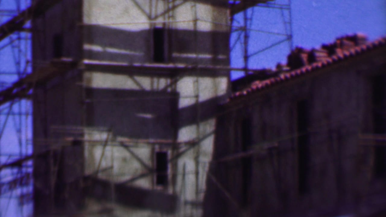 Construction scaffolding crosses a tall pale facade beside a darker roofed building.