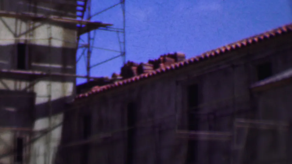 1956 Dallas 8mm frame from a handheld pan across scaffolding, a pale facade edge, and an adjoining masonry building with terracotta roof tiles.
