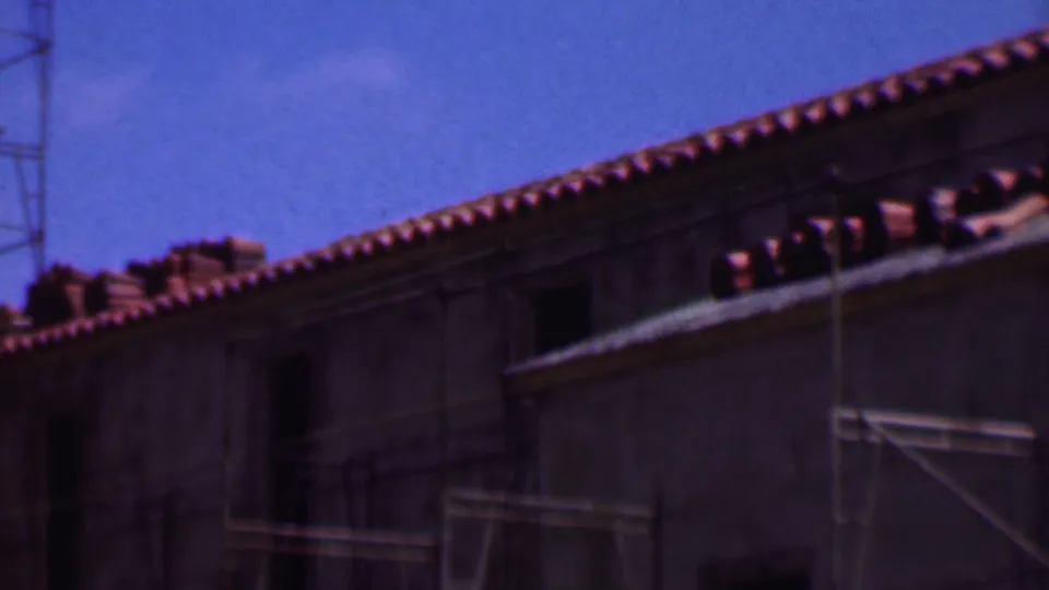 1956 Dallas 8mm frame showing a terracotta tile roofline, dark wall openings, and lower scaffolding beneath a clear blue sky.