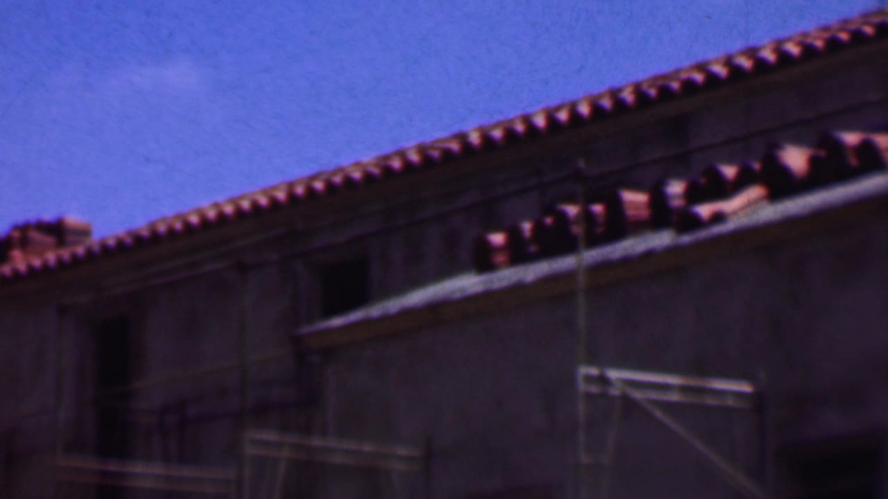 1956 Dallas 8mm frame showing a close view of terracotta roof tiles, a pale building wall, and scaffolding against a bright blue sky.