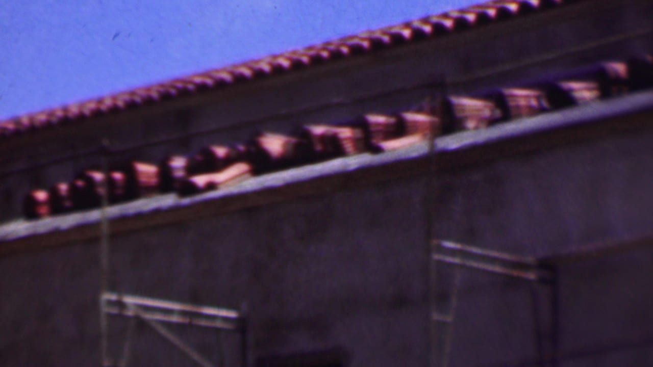 1956 Dallas 8mm frame with curved terracotta tiles along the roofline, a pale wall surface, and metal scaffolding crossing the building exterior.